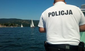 policjant stoi tyłem na białej koszulce napis policja, fragment łodzi policyjnej, w tle jezioro i żaglówki