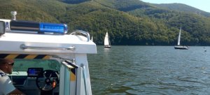 fragment łodzi policyjnej w kabinie siedzi policjant w tle jezioro i żaglówki