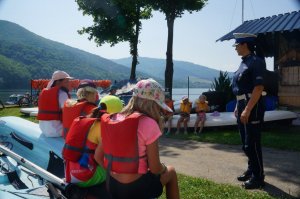 na pierwszym planie tyłem siedzą dzieci w kamizelkach ratunkowych przed nimi stoi policjantka w tle jezioro i góry