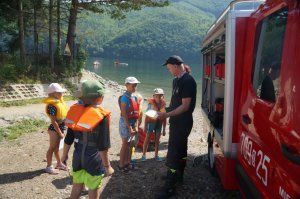 z lewej strony zdjęcia fragment wozu strażackiego obok stoją dzieci i strażak, który pokazuje im sprzęt strażacki w tle góry i jezioro