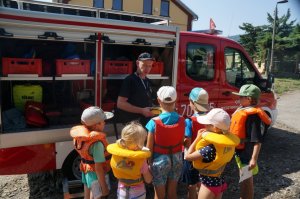 dzieci stoją tyłem przed nimi strażak wszyscy stoją obok wozu strażackiego strażak prezentuje sprzęt strażacki
