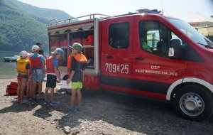 dzieci stoją tyłem obok stoi strażak przed nimi wóz strażacki strażak prezentuje sprzęt strażacki w tle góry i jezioro