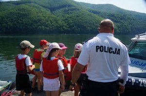 policjant stoi tyłem obok niego na pomoście dzieci wobok policyjna motorówka w tle góry i jezioro
